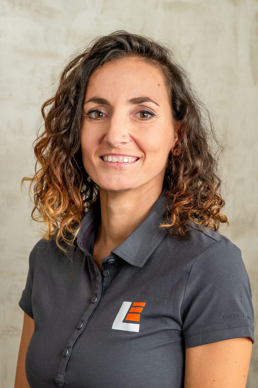 Eine Frau lächelt vor einer grauen Betonwand in die Kamera. Sie trägt ein graues Shirt mit Landeswerk Logo.
